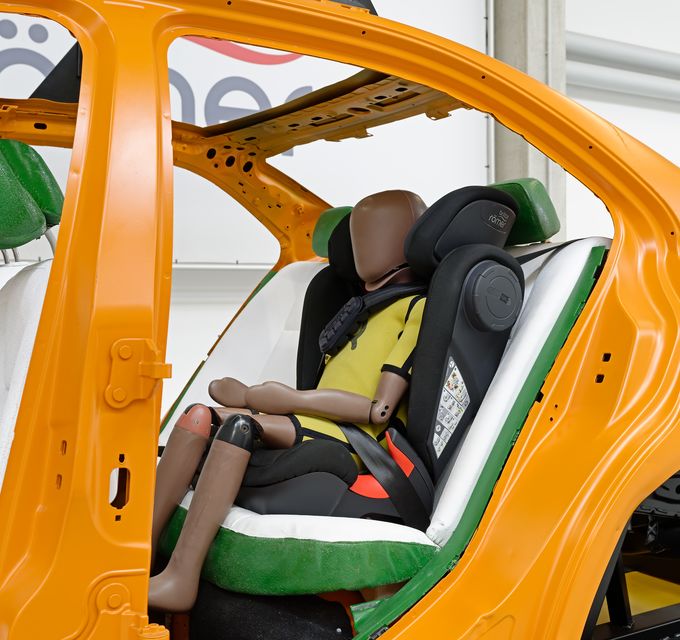 A child crash test dummy is seated in a car seat that is secured to the rear seat of a partially open orange vehicle frame.